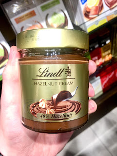 A glass cylindrical jar containing dark brown chocolate spread with bits of gold hazelnut with a gold label with Lindt in gold cursive font with a circular gold plastic lid on a bright background. 