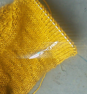 A close up near the live stitches of the sock.  A silver stitch marker in the shape of a feather is locked into the knitting by a lobster clasp.