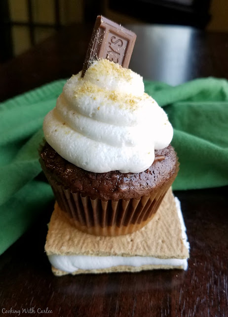 Chocolate cupcakes with a graham cracker crust and piled high with toasted marshmallow buttercream. S'mores cupcakes are a perfect bite of summer! s'mores cupcake perched on top of a graham cracker and toasted marshmallow sandwich