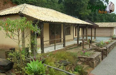 Construção de cabana rústica com a base de pedra, os pilares de madeira no terraço e o piso de cimento queimado branco com a cobertura com telha colonial.