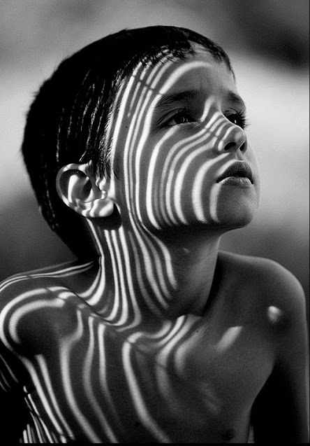 Retrato em preto e branco de um menino olhando para o lado, retrato com luz e sombras formando listras no rosto do menino
