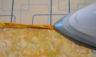 The fabric is on the ironing board. The fabric has been folded once already and is being folded again with the iron on the right hand side to press down the fold.