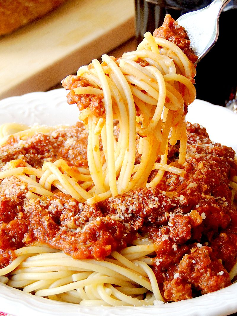 Spaghetti with Slow Cooker Bolognese Sauceon it being twirled on a fork in a white bowl with a red and white checkered napkin on a wooden table with bread and a glass of wine.
