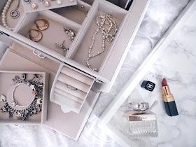 flatlay of jewellery box filled with jewellery next to a tray with perfume and lipstick on it