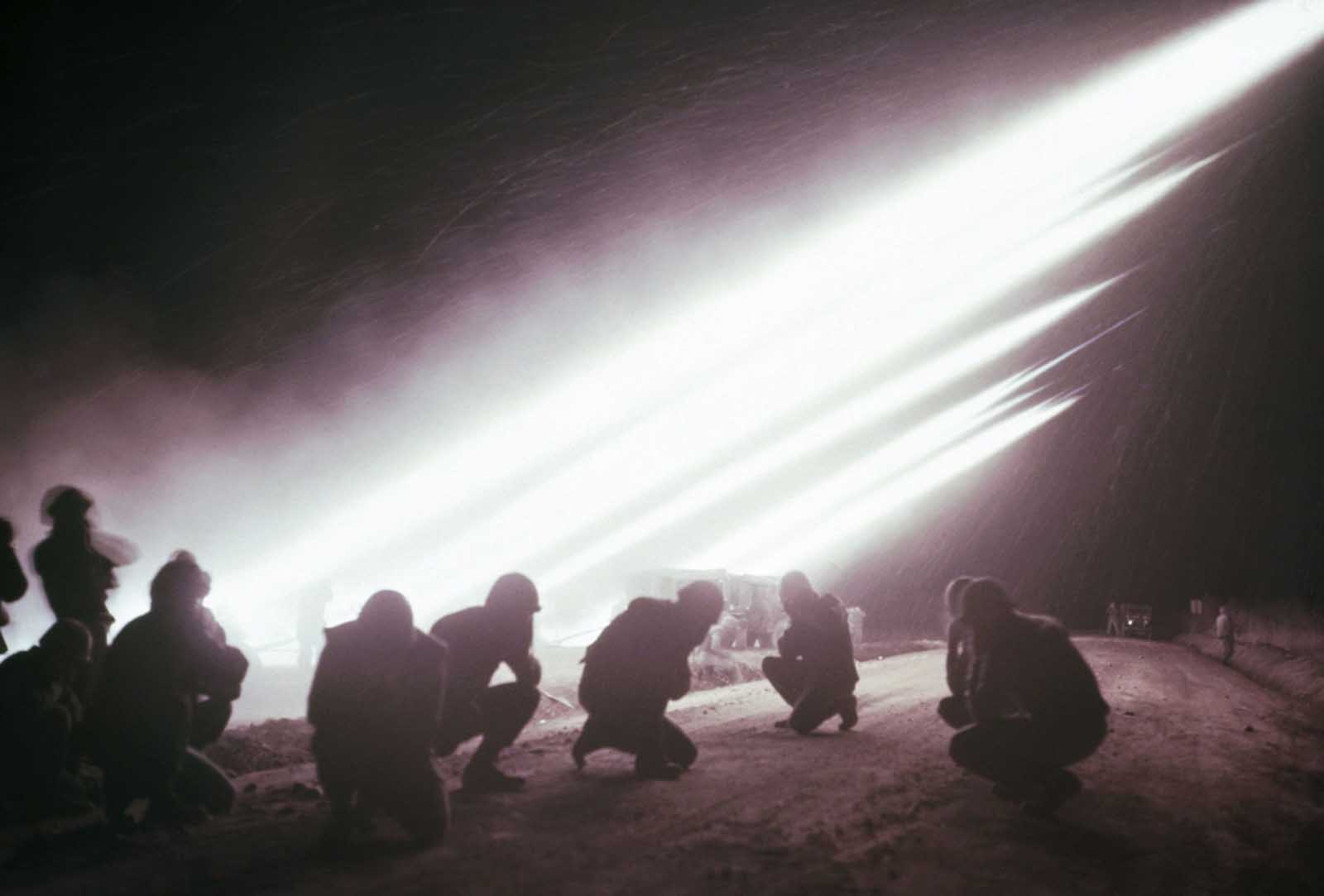 Crew members of a marine battery launcher hold their ears and crouch to the ground as rockets are fired into the night sky.