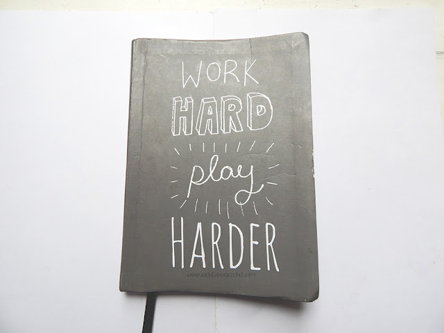 light grey notebook with hand written text on the cover reading "work hard play harder"