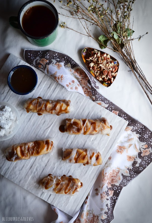 PLANO CENITAL DE ROLLITOS BANOFFEE SOBRE CERÁMICA BLANCA, MANTEL BLANCO, FLORES SECAS, TAZA VERDE DE TÉ Y SALSA CARAMELO