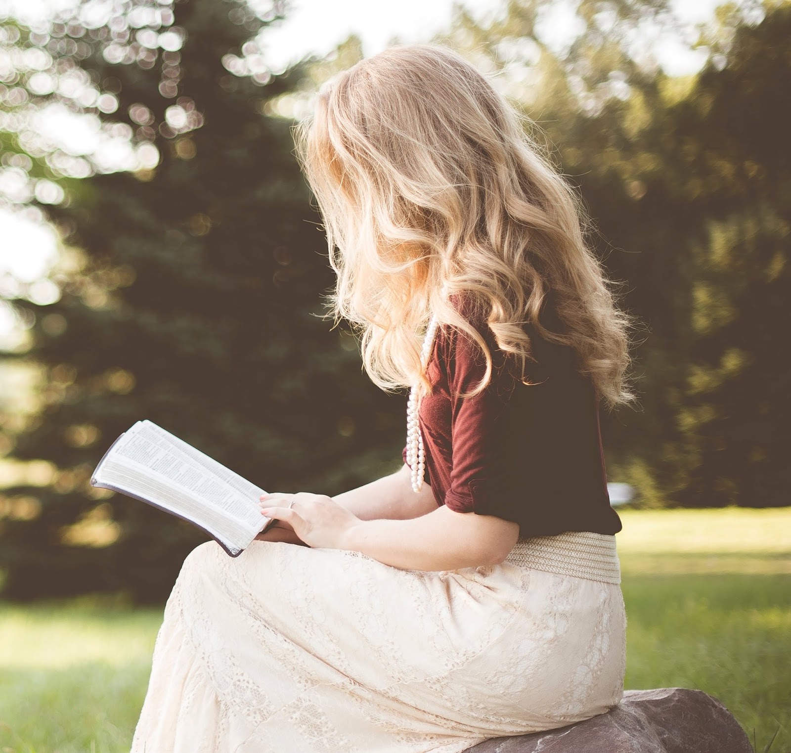 A blond woman sitting on a rock in a park, facing left, reading an book that's on her lap, with trees in the background