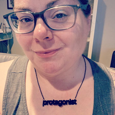 image of me from the shoulders up sitting in my living room with my hair pulled up, wearing grey-framed glasses, a grey tank top, and a necklace that says in black writing 'protagonist'