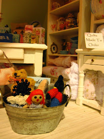 Modern miniature shabby chic shop display of soft toys in a tin tub on the floor, with a display of quilts on cream shelves behind.