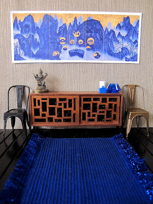 Modern dolls' house miniature scene showing a blue and gold modern chinese panel on the wall. Underneath is a sideboard with a variety of blue and gold items on it, and beside it, two french cafe chairs in black and gold.