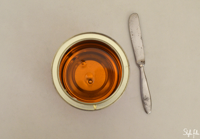 A flatlay image of honey like hot wax in a metal tin along with a metal applicator on a white background