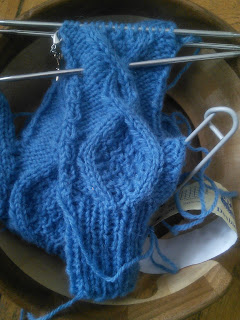 A knit mitten with cables done in blue yarn. The mitten is still live on double-pointed needles. The thumb gusset stitches are on a stitch holder. Mitten and yarn are tucked into a wooden yarn bowl.