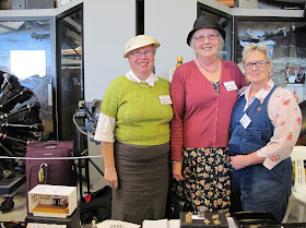 Three women dressed in 1940s outfits behind a stall at a miniatures convention.