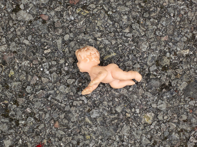 Tiny, naked, pink, plastic doll on a tarmac path.