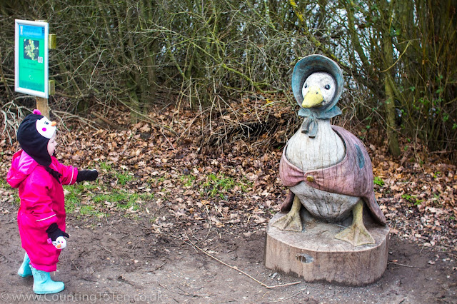 A toddler pointing at a wooden carving of Beatrix potter's Jemima Puddleduck next to a lake