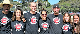 Group of camp counslors wearing their staff t-shirts and standing together on the beach with their arms around each other.