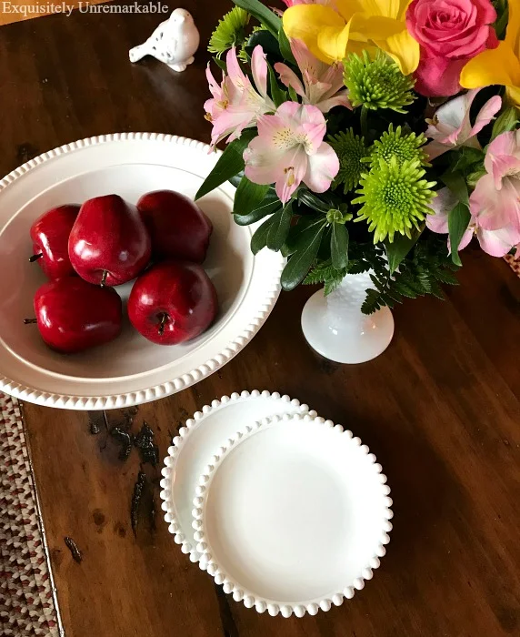 Faux white milk glass dishes, bowl filled with apples and a white flower filled vase on coffee table