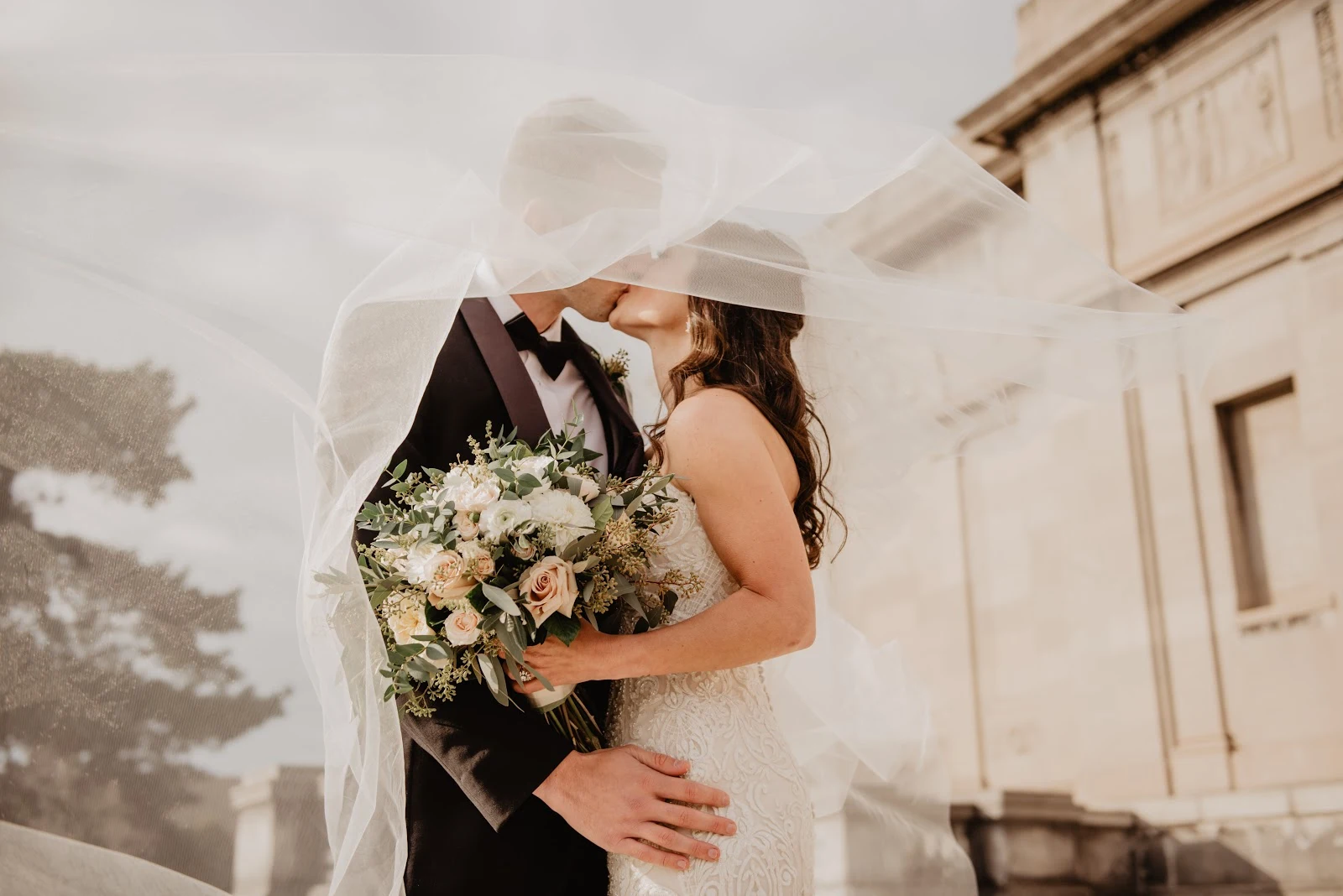 結婚式で口付けを交わす黒いタキシードの男と白いドレスの女に白いベールがかかっている