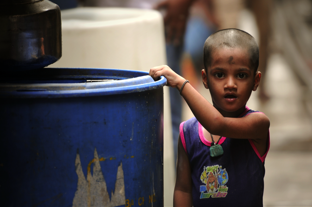 In this picture a girl is standing next to a blue water barrel in an alley in Dharavi, India. The picture is just one out of many of the shadow city of Mumbai taken by the photographer.