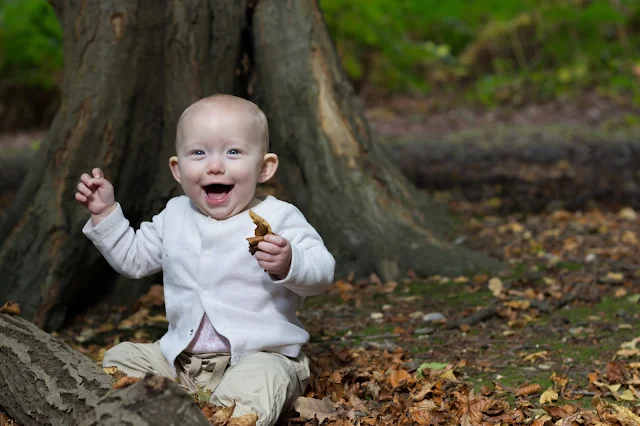A 9 month old baby sitting on the floor if the forest on brown leaves next to a log and in front of a tree