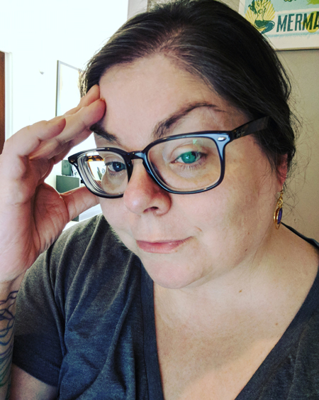 image of me at my desk, with my hair pulled back, wearing grey-framed glasses and a grey t-shirt and gold earrings with small pieces of purple agate