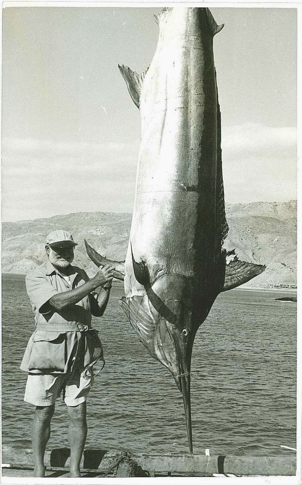 A black and white photograph of Hemingway next to a hanging marlin.