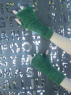 Someone wearing a pair of fingerless mittens. The mittens are knit in a dark green fingering-yarn and use a textured star stitch over the body of the mitten