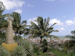 Links ein Denkmal aus Beton des Hatuey, zwischen Palmen und grüner Vegetation hoch oben über der Stadt Baracoa und dem Meer. Kuba, Baracoa, Statue es Hatuey über Stadt und Bucht von Baracoa