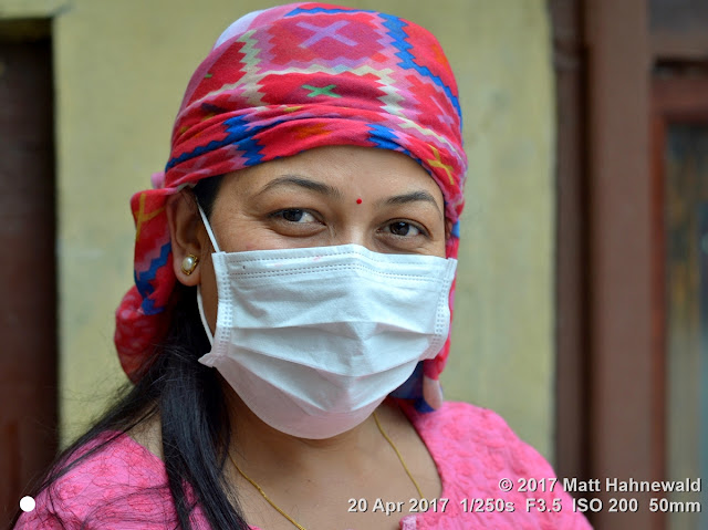 Closeup street portrait (outdoor headshot, three-quarter view) of a mature Nepali woman with beautiful eyes, wearing a disposable surgical earlobe face mask; Kathmandu, Nepal. Matt Hahnewald Photography; Facing the World; photography; photo; image; amazing; spectacular; lively; outstanding; fantastic; favourite; superior; interesting; Nikon D3100; Nikkor AF-S 50mm f/1.8G; prime lens; 50mm lens; 4 : 3 aspect ratio; horizontal format; street; portraiture; portrait; closeup; headshot; three-quarter view; pink; outdoor; colour; colourful; character; personality; real people; human head; forehead; bindi; human face; human eyes; talkative eyes; smiling eyes; eye contact; face mask; headscarf; disposable surgical mask; enigmatic; sexy eyes; hygiene; consent; empathy; rapport; encounter; emotion; ethnic portrait; travel portrait; Hinduism; Kathmandu; Nepal; mature woman; Nepali woman; one person; female; beauty; adult; posing; authentic; smiling; smizing; fabulous; attractive; sensual; soulful; captivating; charming