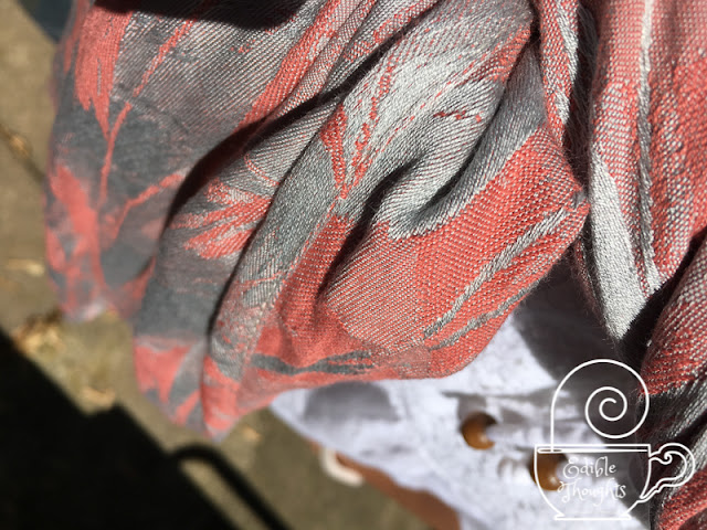 [Close-up of a silver and coral painterly patterned woven wrap in the sunlight. The silver threads shimmer in the light.]