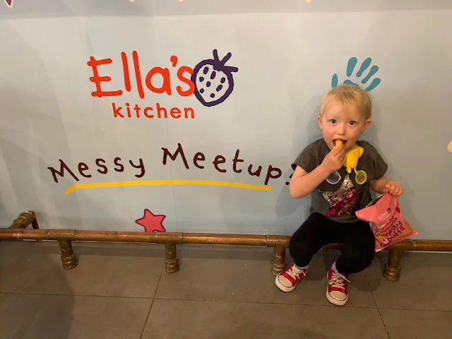 A poster saying "Ella's Kitchen Messy Meetups" and a toddler eating some food witha toy banana stuffed down her t-shirt"