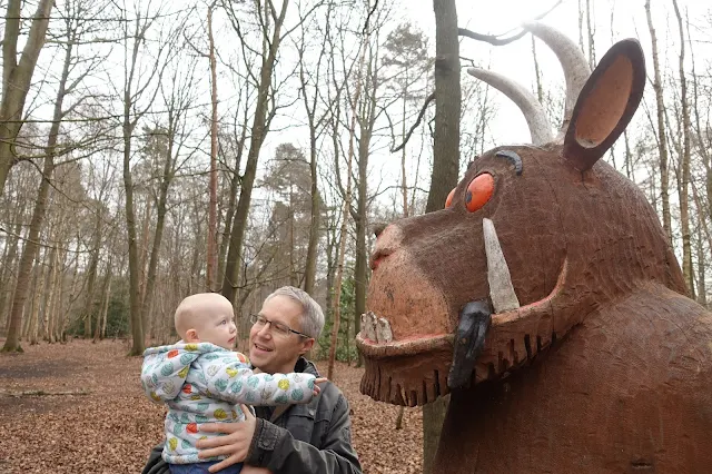 A baby held by her daddy is looking at a carved gruffalo with a black tongue