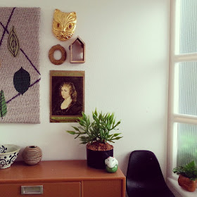 One-twelfth scale modern miniature office scene containing a credenza with a black Eames chair next to it and an artwork with leaves above it. On the credenza is a bowl, a vase, a potted plant and a ceramic ornament. On the windowsill to the right is a potted plant.