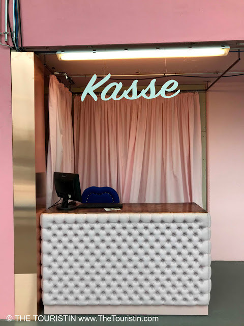 A ticket booth decorated with soft pink fabric, sporting the letters Kasse in light green above the screen of the register.