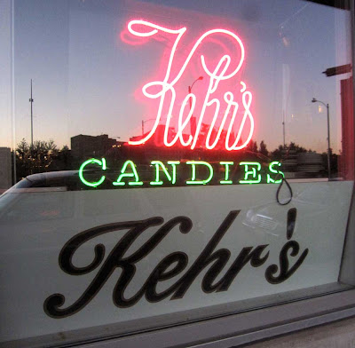 Photo of a store window with lit neon reading Kehr's Candies and printed sign below saying Kehr's with the apostrophe way over above the letter s