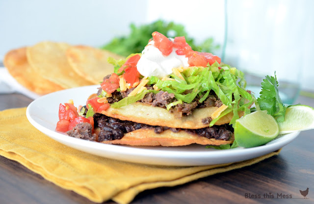 A beef tostada on a white plate with cilantro, lime, and fried tortillas sitting by the plate.