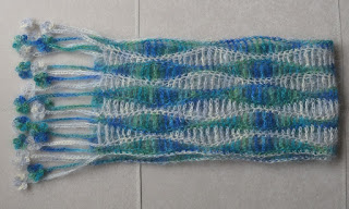 Folded silk mohair scarf. Fold is on right hand side; floral fringe is on left hand side. The scarf has a lengthways wavy stripe pattern alternating in blue-greens and silver-greys.