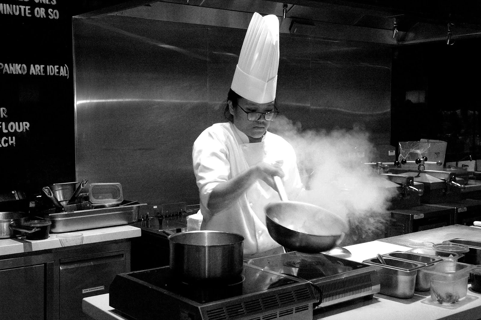 A chef in a professional kitchen, intensely focused on tasting a sauce with a spoon.