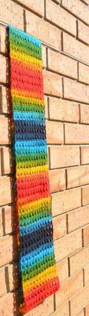 Side view of cowl hanging flat against a light orange brick wall. It is approximately 10 brick-heights long. The cowl is in rainbow stripes.