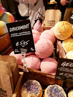 A group of bright pastel green spherical bath bombs on a large wide rectangular light brown shelf with a black board saying Chocomint in white font on a bright background 