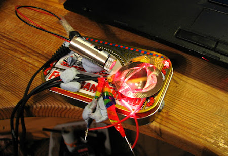 [Image: A mess of wires and components attached to a mint tin using blu-tack. A laser module is pointing at a piezzo buzzer at an acute angle. A piece of mirror is glued onto the piezo buzzer. The laser and piezzo buzzer are connected to wires that lead out of the picture.]