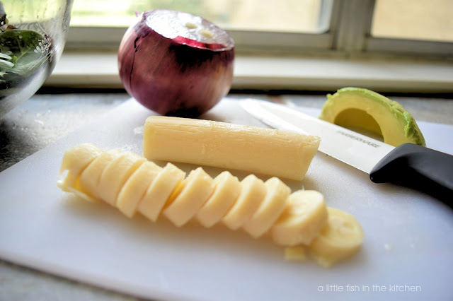 A heart of palm is sliced and ready to use on a white cutting board. A sliced sections of green avocado and one half of a purple onion  are beside a knife on the cutting board also. 