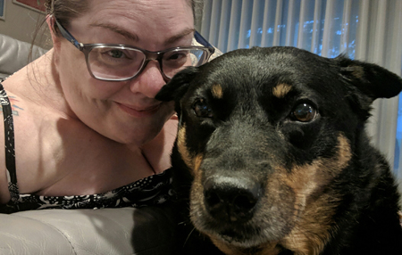 image of me on the couch, wearing a strappy sleeveless top and grey-framed glasses, with my hair pulled back, smiling and posing next to Zelda the Black and Tan Mutt, who is sitting in front of me in a protective position
