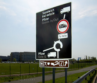 Photograph of a road sign with a black background and white text and symbols. There is als a brown information sign underneath attached to the same poles showing white coloured service symbols.