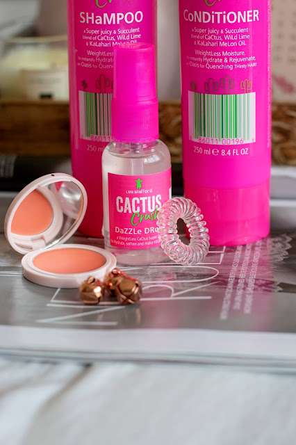 Close up Lee Stafford Cactus Crush shampoo and conditioner, pink bottles. Cactus Crush dazzle drops, invisibobble and peach cream blusher on magazine with July calendar on tray in background with flowers in blush pink vase