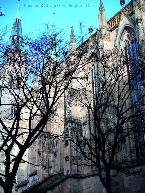 Photo of the St. Sebald Church in Nuremberg,  Germany. Photo shows the side wall of stone with towering windows showing in the sunshine.  To the left is one of the Gothic towers on the church with  blue spire. by RosevineCottageGirls.com