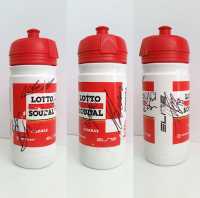 A collage of three different views of the same plastic bidon. It is white with a red top, with sponsors' names printed in red, white and black. The team name is in large black block letters "Lotto Soudal" on a white backkground, with a red thick strip between the words and surrounded by solid red background. Underneath the team name are thin strips of black yellow and red (Belgian flag colours). In the red background are names of secondary sponsors in white writing. The white strip along the side has the word "Elite" printed lengthways along the bidon in black.  Signatures are written in black texta.