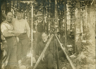A black and white photograph of men and surveying equipment in a wooded area.