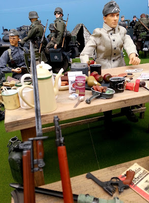 1/6 scale German soldier sitting at a table with food on it in diorama of an army post on display at a scale model exhibition.